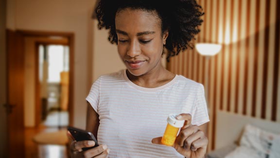 An African American woman is sitting on the bed in the bedroom communicating with the doctor online while holding a bottle of medicine in her hand